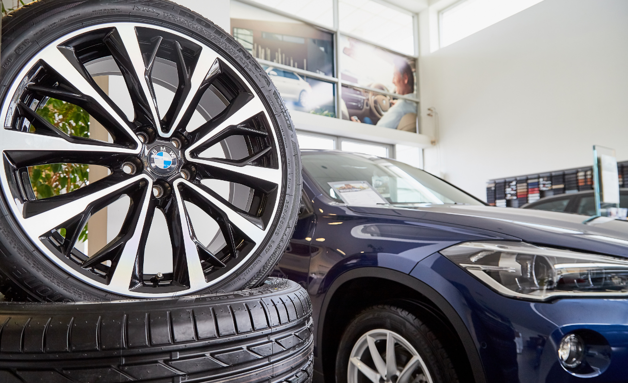 A close-up of a BMW alloy wheel is displayed on stacked tires in the foreground. In the background, a blue BMW SUV is parked indoors, partially visible with showroom decor and a large window. EuroMechanic | Redline Automotive | Top Reviewed Auto Repair Shop in Scarborough | Visit redlineautomotive.ca
