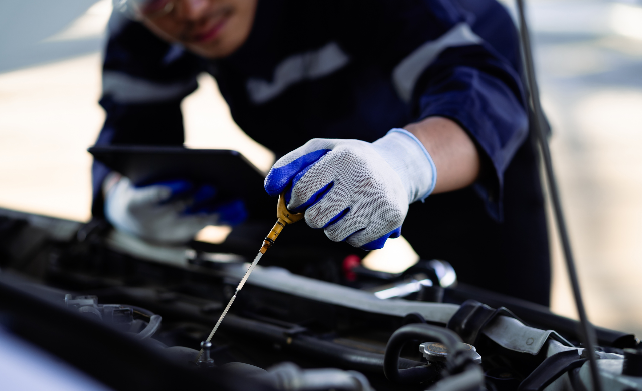 A mechanic in a dark uniform and white gloves inspects a car engine. They are holding an oil dipstick, checking the oil level, while using a digital tablet. The focus is on the engine and dipstick, with the mechanics face partially visible. EuroMechanic | Redline Automotive | Top Reviewed Auto Repair Shop in Scarborough | Visit redlineautomotive.ca
