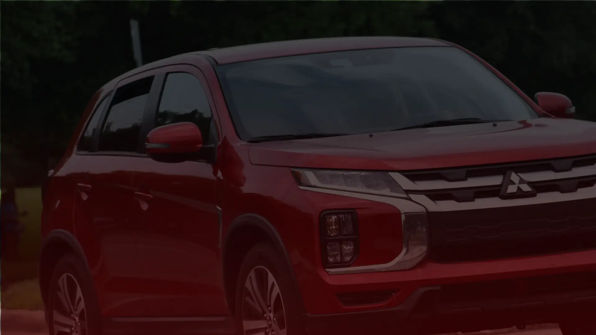 A red Mitsubishi SUV is parked outdoors, its sleek design accented by modern headlights and prominent branding on the grille. In the distance, greenery frames Scarboroughs natural scenery under a cloudy sky, hinting at the precision of a Mitsubishi mechanics craftsmanship. EuroMechanic | Redline Automotive | Top Reviewed Auto Repair Shop in Scarborough | Visit redlineautomotive.ca