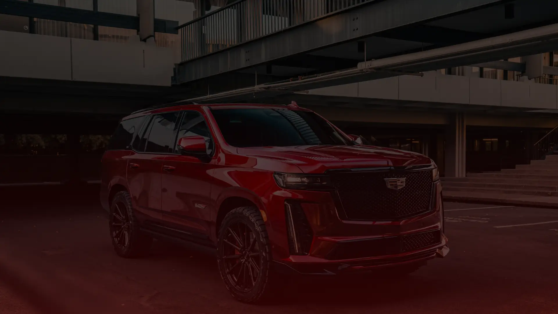A shiny red Cadillac SUV, likely serviced by a skilled Cadillac mechanic in Scarborough, is parked in an urban setting with modern architecture. Its sleek design, large black wheels, and tinted windows create a striking contrast against the surroundings. EuroMechanic | Redline Automotive | Top Reviewed Auto Repair Shop in Scarborough | Visit redlineautomotive.ca