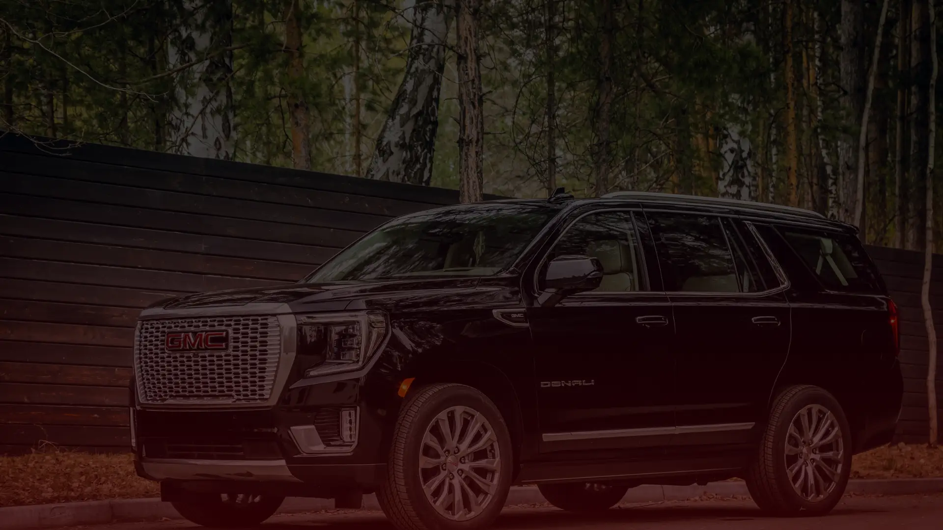 A black GMC Denali SUV is parked on a road, framed by a tall wooden fence and lush forest. The vehicles distinctive grille and sleek design catch the eye, showcasing its robust style—ideal for anyone seeking quality craftsmanship from a GM mechanic in Scarborough. EuroMechanic | Redline Automotive | Top Reviewed Auto Repair Shop in Scarborough | Visit redlineautomotive.ca