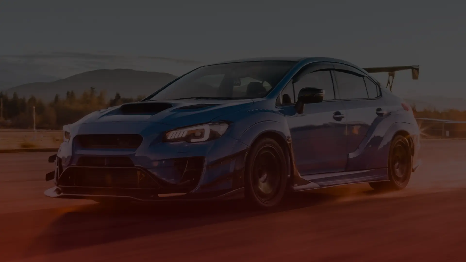A blue sports car with a large rear wing, tuned by a skilled Subaru mechanic in Scarborough, speeds down the track. Its aerodynamic design and aggressive stance are highlighted against the blurred backdrop of mountains and a clear sky at dusk. EuroMechanic | Redline Automotive | Top Reviewed Auto Repair Shop in Scarborough | Visit redlineautomotive.ca