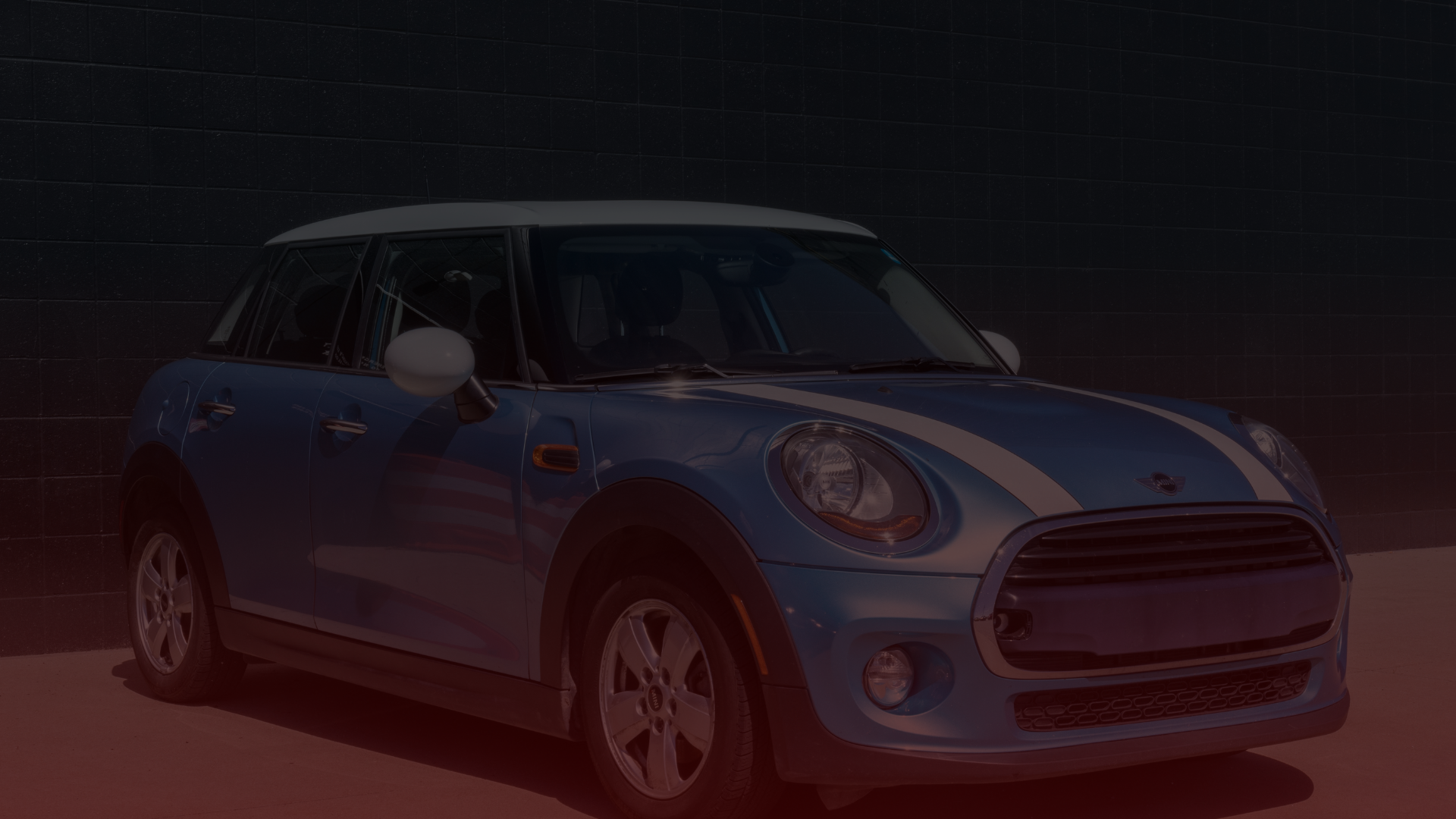 A blue Mini Cooper with white racing stripes is parked in front of a dark brick wall. Sporting a white roof, side mirrors, and chrome accents, its perfectly positioned on sunlit pavement. If you need a Mini Cooper mechanic nearby, check out the top Mini Cooper repair shop in Scarborough. EuroMechanic | Redline Automotive | Top Reviewed Auto Repair Shop in Scarborough | Visit redlineautomotive.ca