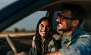 A man and woman are sitting in a car, both smiling and looking happy. The woman has long dark hair, and the man is wearing sunglasses and a denim jacket. Sunlight is streaming through the window, suggesting a sunny day outside. EuroMechanic | Redline Automotive | Top Reviewed Auto Repair Shop in Scarborough | Visit redlineautomotive.ca