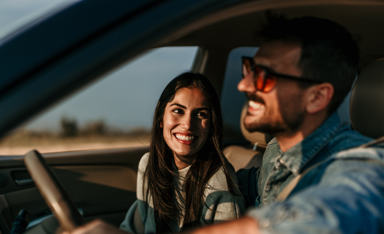 A man and woman are sitting in a car, both smiling and looking happy. The woman has long dark hair, and the man is wearing sunglasses and a denim jacket. Sunlight is streaming through the window, suggesting a sunny day outside. EuroMechanic | Redline Automotive | Top Reviewed Auto Repair Shop in Scarborough | Visit redlineautomotive.ca