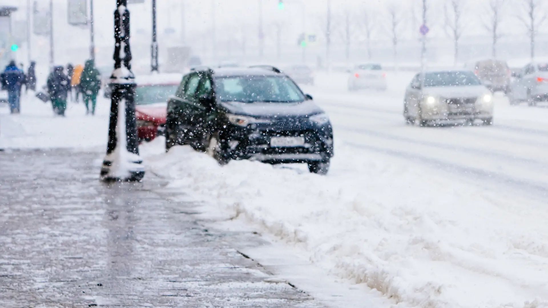 Cars drive down a snow-covered street during heavy snowfall. A sidewalk runs along the left, lined with snow piles, and a few people walk bundled up in winter clothes. Visibility is reduced due to the falling snow. EuroMechanic - Redline Automotive | Top Reviewed Auto Repair Shop in Scarborough | Visit EuroMechanic - Redline Automotive
